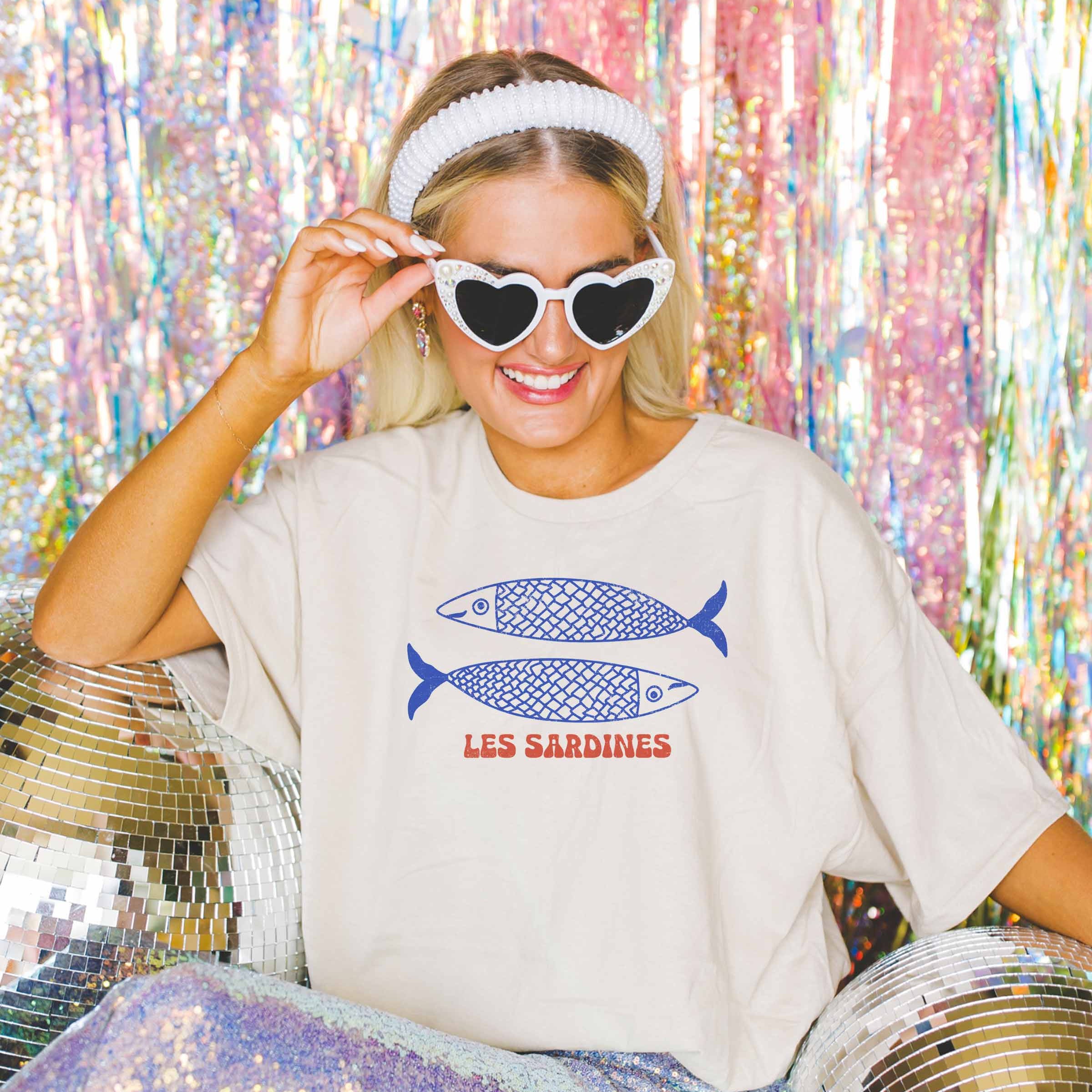 Frau trägt ein Oversized T-Shirt Les Sardines mit Grafikdruck, umgeben von Discokugeln und Glitzerhintergrund.